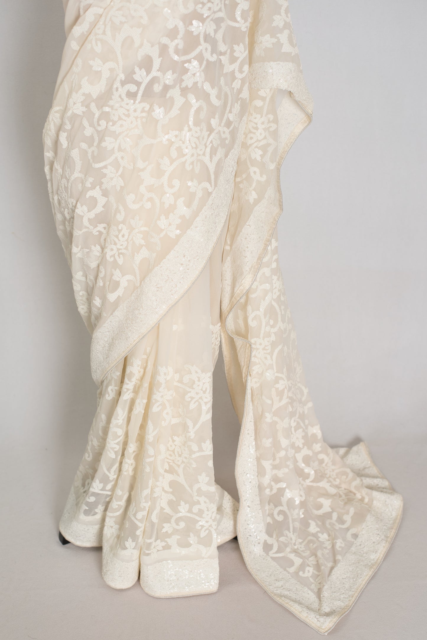 An ivory/cream saree with intricate floral lace embroidery, draped elegantly to showcase its texture and pattern against a neutral light gray background.