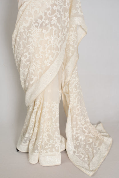 An ivory/cream saree with intricate floral lace embroidery, draped elegantly to showcase its texture and pattern against a neutral light gray background.