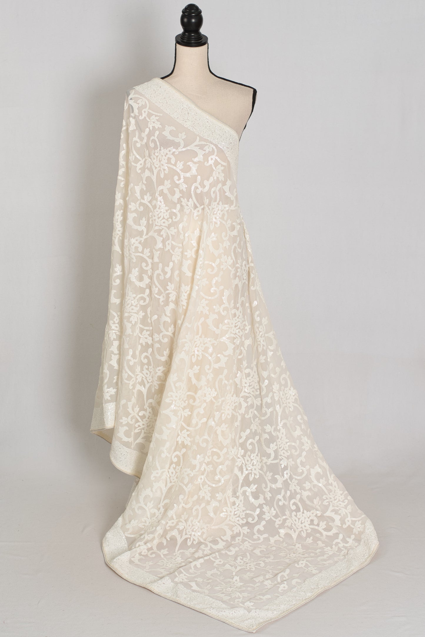 An ivory/cream saree with intricate floral lace embroidery, draped elegantly to showcase its texture and pattern against a neutral light gray background.