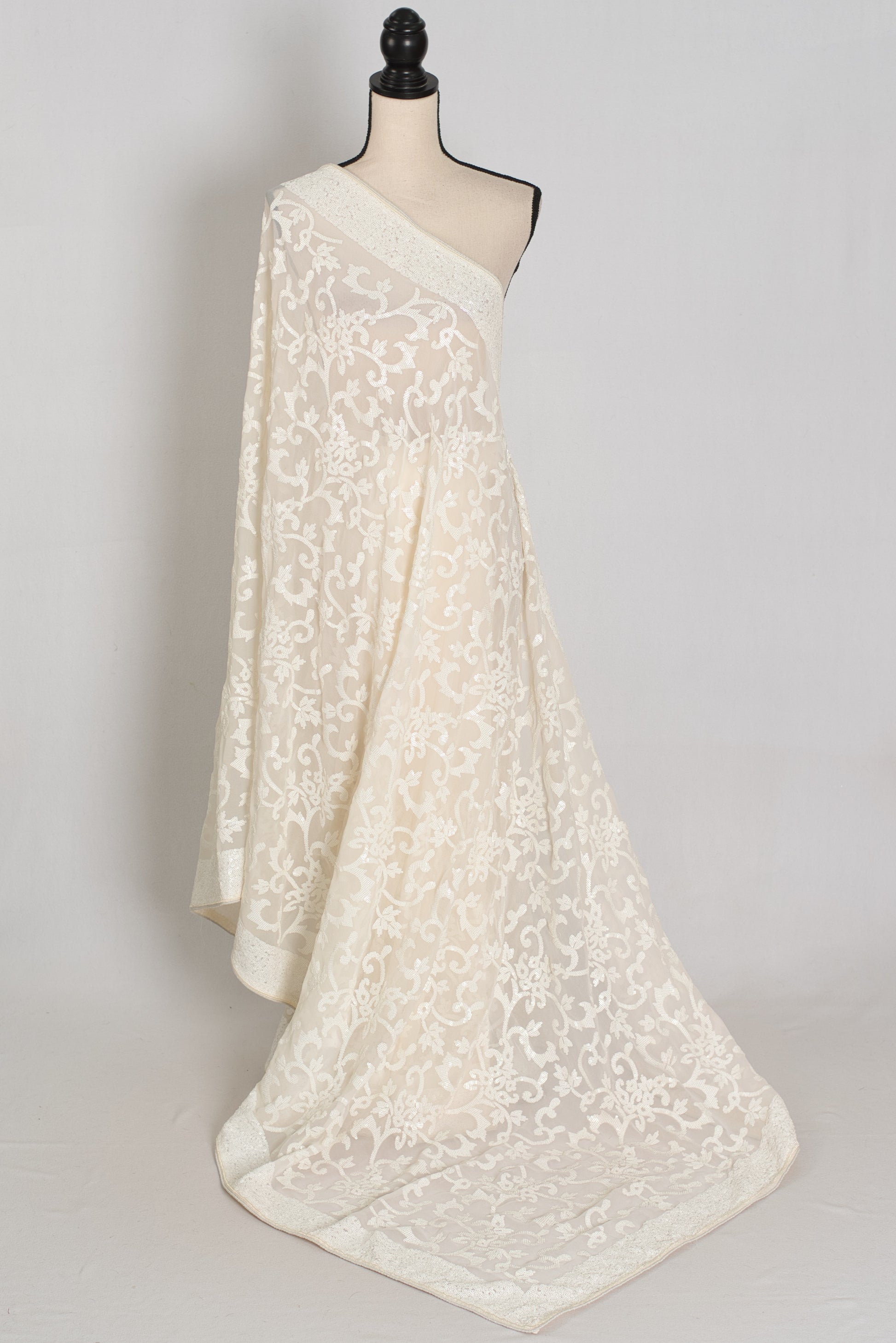 An ivory/cream saree with intricate floral lace embroidery, draped elegantly to showcase its texture and pattern against a neutral light gray background.