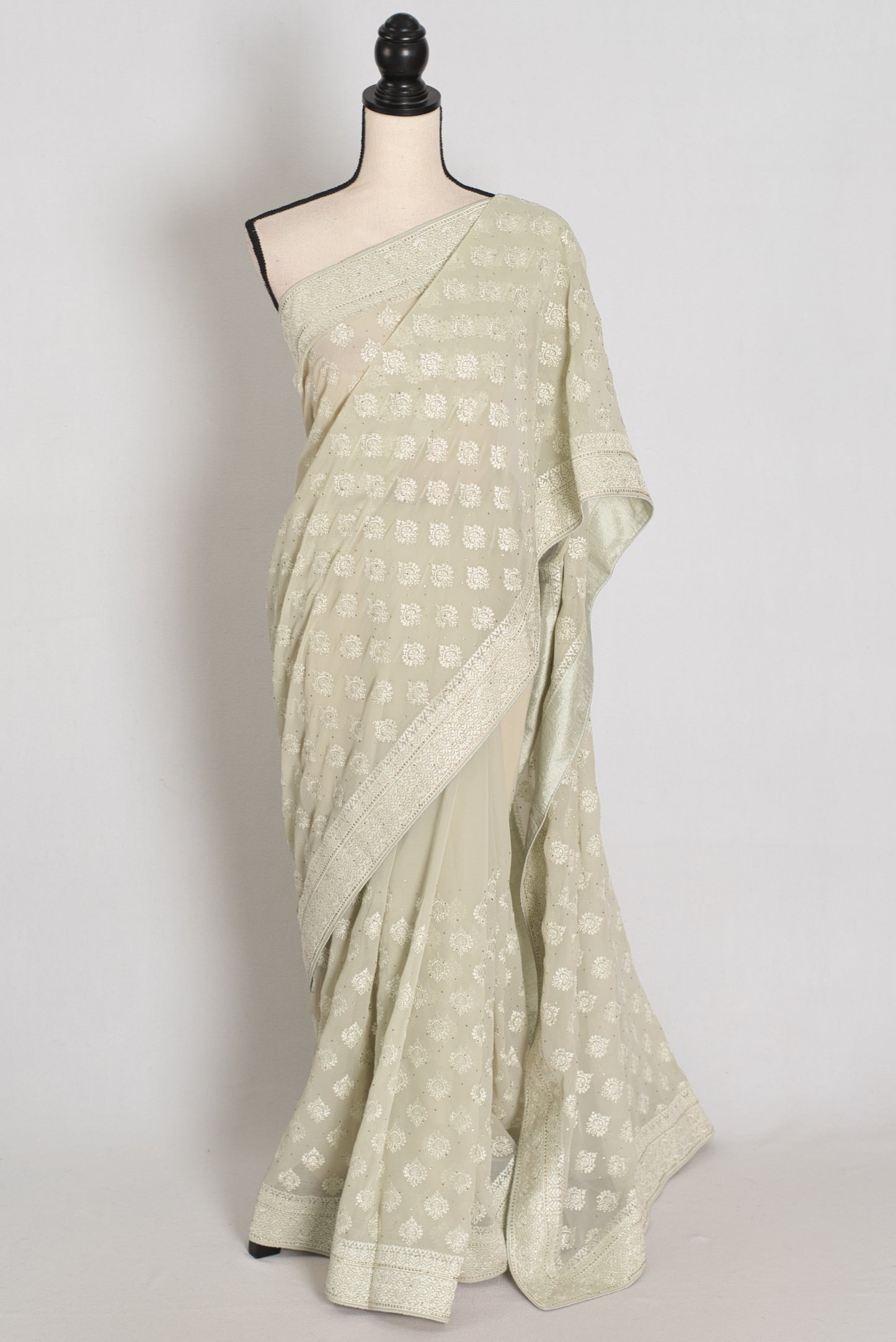 A light sage green saree with a white geometric pattern and a wide border, displayed on a mannequin against a plain light gray background.